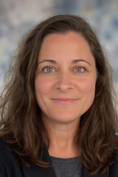 A white woman with long brown hair, blue eyes, and a grey sweater stands in front of a grey background.