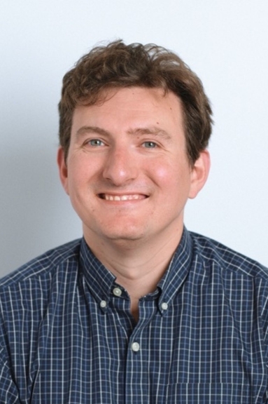 A man, with a blue plaid button-down shirt, and short brown hair, smiles into the camera.