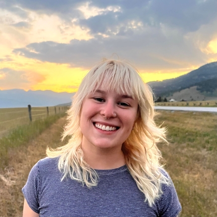 A smiling person with past shoulder length blonde hair in front of a mountainous background
