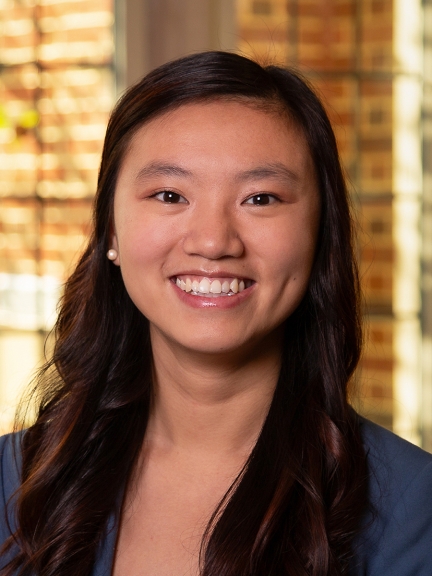 Headshot of Mallory Haskins - woman with long dark-brown curled hair and brown eyes smiling warmly at the camera wearing a navy blazer and pearl earrings