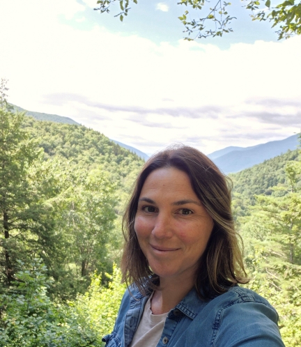 A smiling person with shoulder length hair in front of a mountainous background