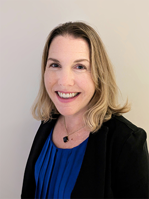Headshot of Brooke Fox, wearing black blazer over royal blue pleated top with blue necklace