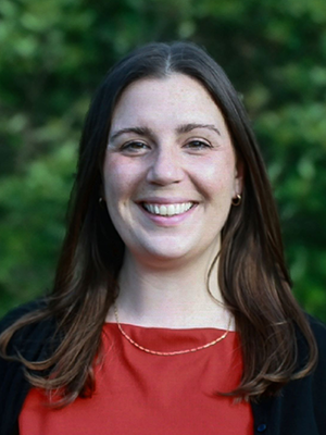 headshot of Caroline Santoro in muted red blouse and black blazer against a backdrop of greenery
