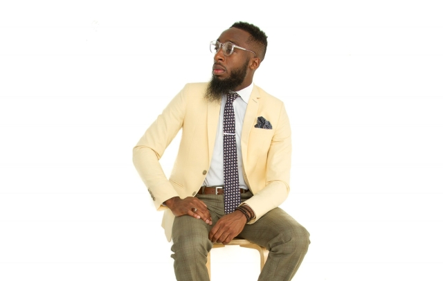 Blackm man wearing a yellow suit jacket seated in a stool, looking to the side