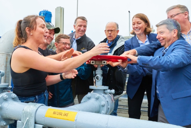 A group of smiling white people in blue and black clothing turn the wheel to start a manure digester 