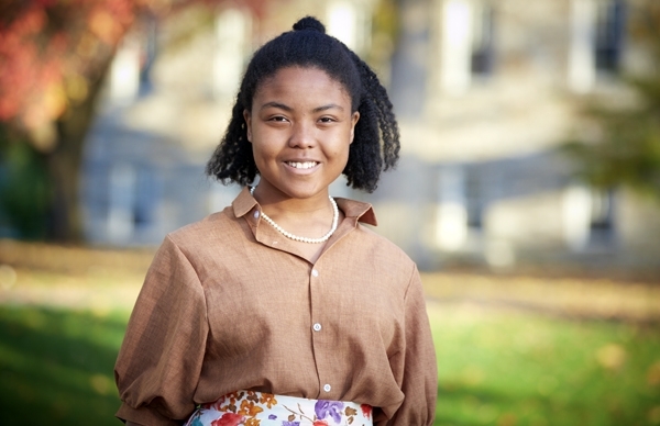 Christine McDow smiles at the camera in pearls, a brown blouse, and a multi-colored skirt. 