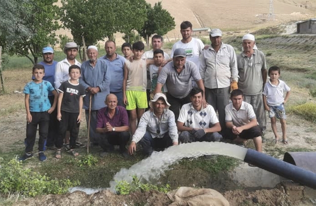 A group of villagers pose for a photo behind a rush of water spewing from a newly constructed irrigation channel