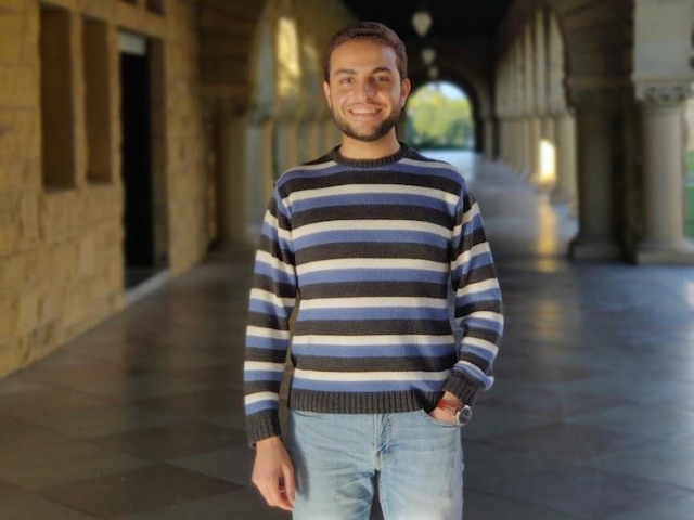 Osama Abdelrahman poses in a long-sleeve shirt designed with horizontal blue, white, and gray lines. He stands in front of a long hallway.