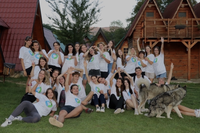 A group of young people, all wearing the same white t-shirt with a green and blue emblem, pose for a silly group photo in front of a log cabin.