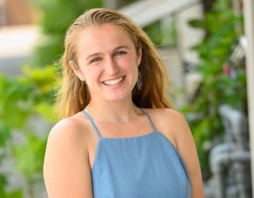 Allie Stankewich poses smiling for a photo in front of a green garden background while wearing a blue, sleeveless, blouse.