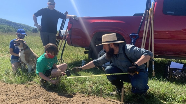 Pete Antos-Ketchum makes preparations in soil of a garden bed, one child in a face masks helps him. Another child plays with golden retriever while another man leans against a red truck watching them work. Foreground of mountains and blues skies. 