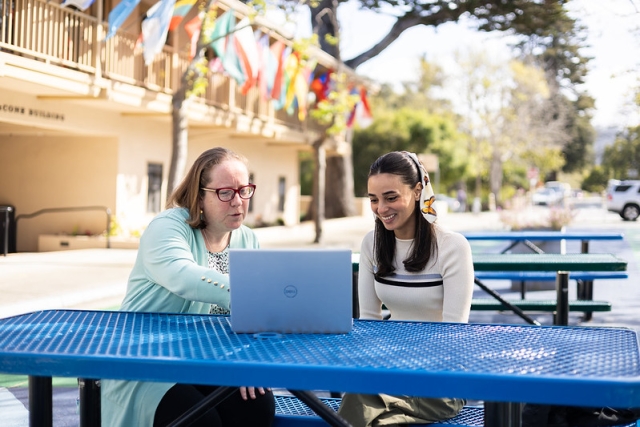 two women sit at a picnic table with a laptop open in front of them