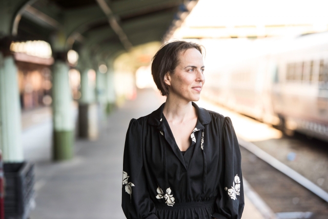 A woman with short hair looks away from the camera while standing on a train platform
