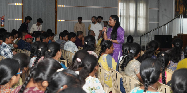 A person wearing a purple dress speaks to a room of people. 