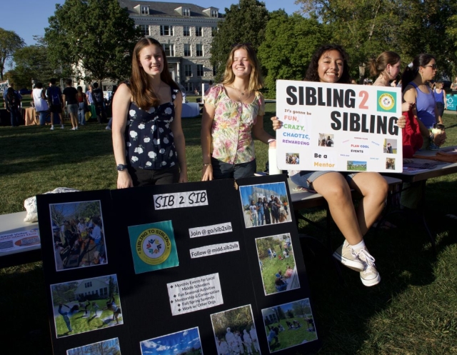 Three students smiling standing outside at a tabling event with signs and posters promoting Sib2Sib