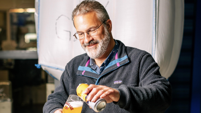 Rob Tod stands in front of a bag of hops and smiles while pouring an Allagash White beer from a can into a glass.