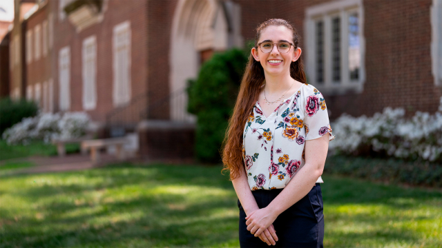 Person standing on college campus smiles and looks at the camera.
