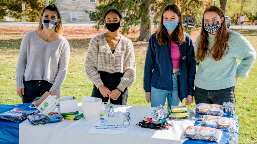 Four Middlebury students stand, wearing masks, around an outdoor table laden with masks to give away.