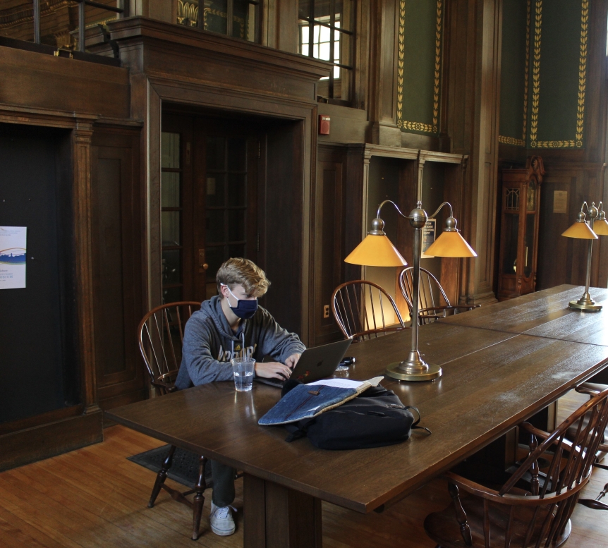 A student wearing a mask works on his laptop atop a large wooden table.