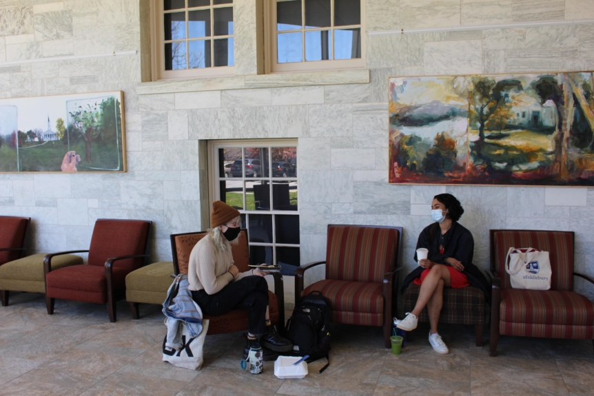 Two students wearing masks, sit distanced and chatting in Axinn. Large art pieces hang on the wall behind them.