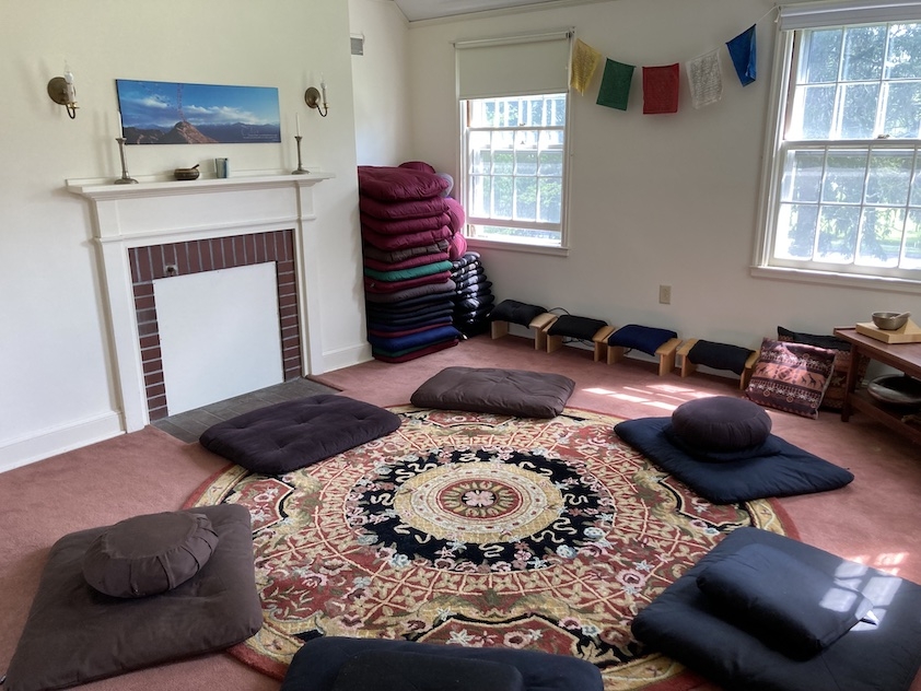 Meditation room with cushions, a circular rug, and prayer flags. There are windows letting in sunlight and the room has a soft pinkish red color scheme.  