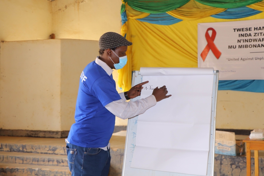 A man in a cap, mask, and blue t-shirt writes on a large pad of paper propped up on an easel.