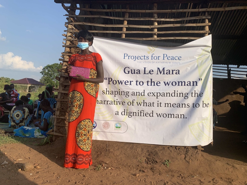 A woman in a mask and orange dress holds a pink woven purse, and stands in front of a white sign with the black text "Gua le Mara Power to the Women"