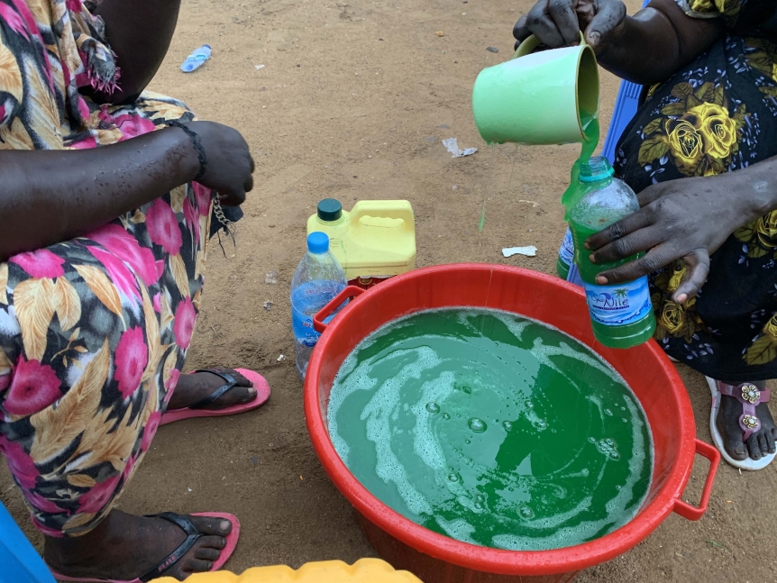 Two pairs of hands scoop green dyed water out of an orange bowl on the sandy ground.