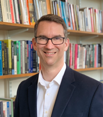 Erick Bleich standing in front of a bok case in dark jacket with glasses