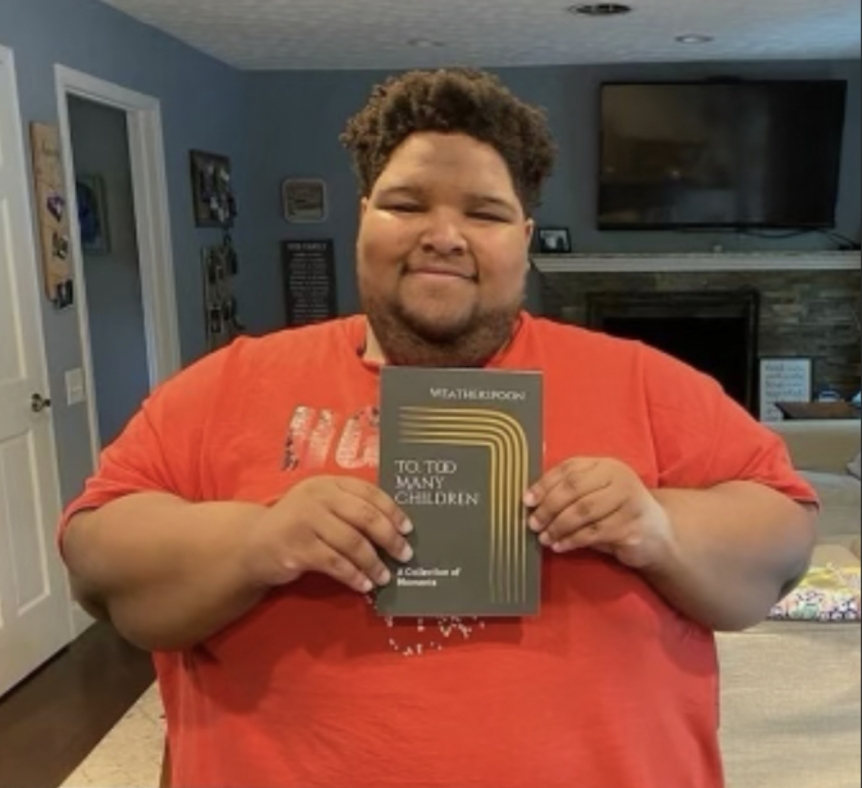 Weatherspoon, wearing an orange t-shirt, poses in a dimly lit living room with their book of poetry held in their hands. 