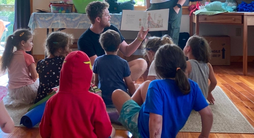 A student is sitting in front of a group of young children reading a book. The young students are all sitting on the floor listening.