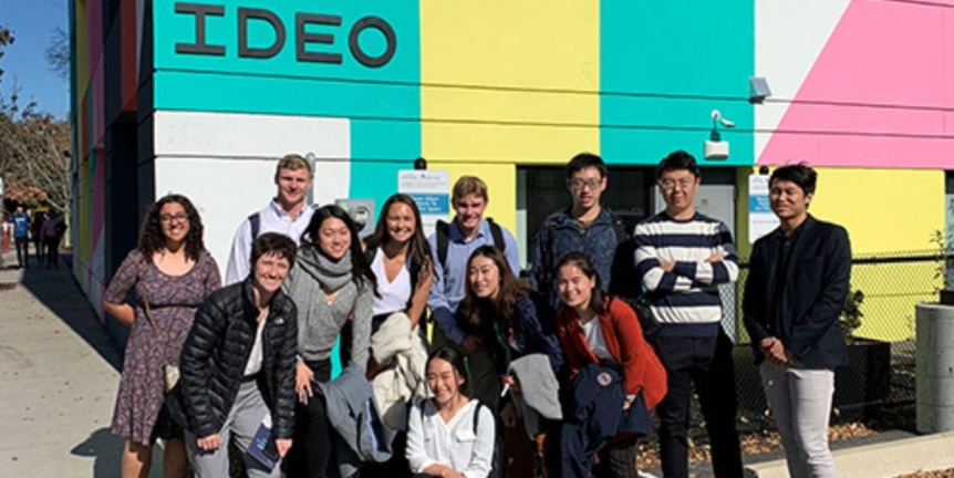 Middlebury students and alumni stanging outside the IDEO headquarters in Boston, MA. Brightly colored IDEO building is behind them.