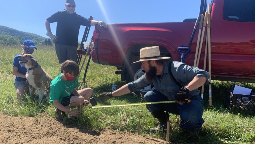 Pete Antos-Ketchum makes preparations in soil of a garden bed, one child in a face masks helps him. Another child plays with golden retriever while another man leans against a red truck watching them work. Foreground of mountains and blues skies. 