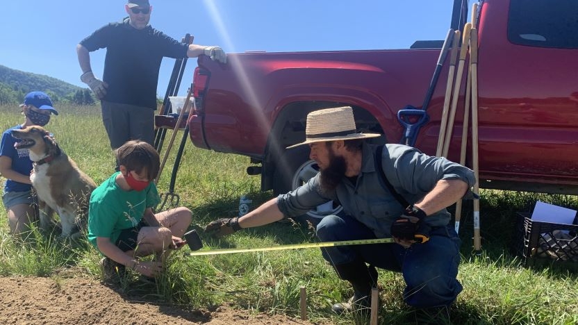 Pete Antos-Ketcham prepares garden bed with help from child in a face mask. A boy with a dog, and a man leaning on a red pickup truck in background.