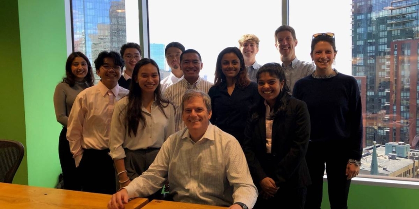 A group of students pose standing behind an alumni in a conference room.