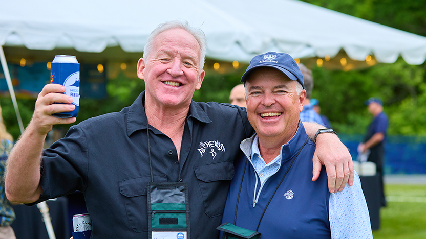 Two friends from 1984 with their arms around each others' shoulders. One raises a can in a toast.