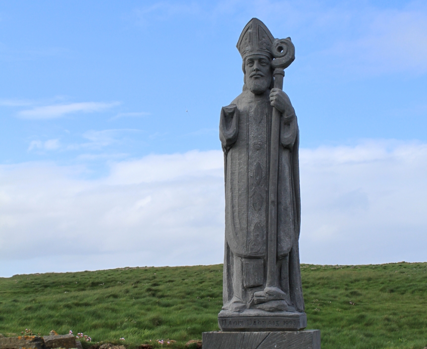 stone sculpture of St. Patrick standing in green field against blue sky