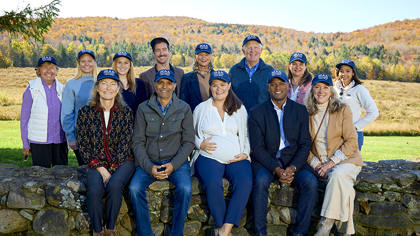 Board members of the Middlebury Alumni Association outdoors on the Breadloaf Campus at a 2024 fall retreat. They are all wearing navy baseball caps saying "MAA."