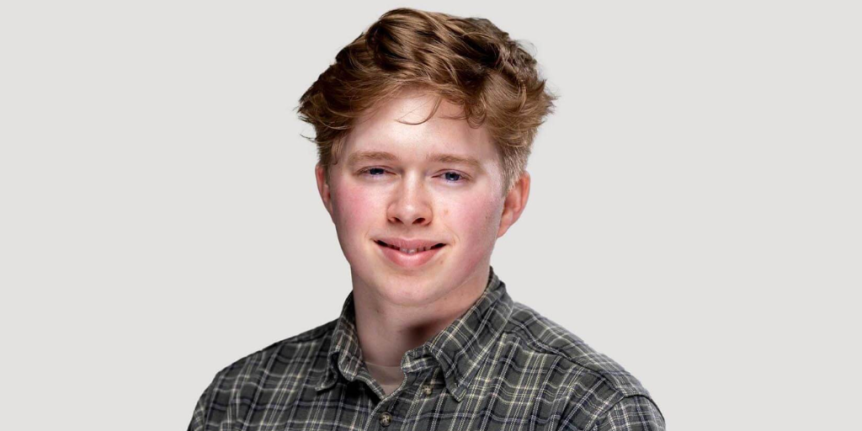 Dominic Needham stands against a white backdrop wearing a plaid shirt.