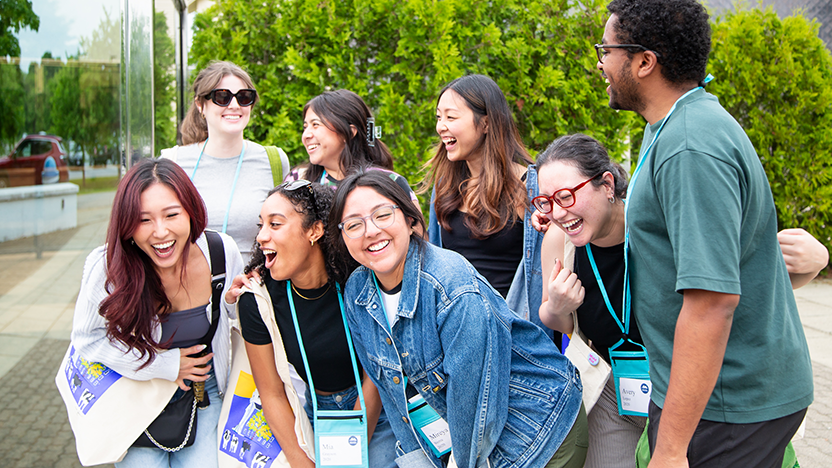 A group of 8 friends from the class of 2020 laughing while arranging themselves for a group photo. Several of them carry Middlebury tote bags featuring a cow and chapel painted by Woody Jackson.