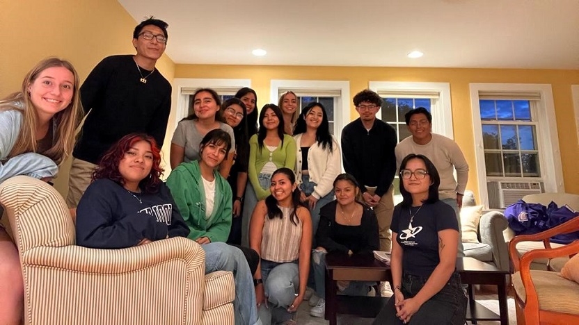 A group of students stand, crouch, and sit on couches, all smiling at the camera.