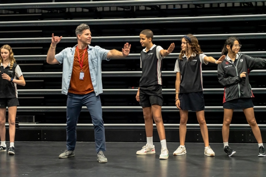 a tall man demonstrates a movement in a gym setting, with four high school students stand around him also doing the movement