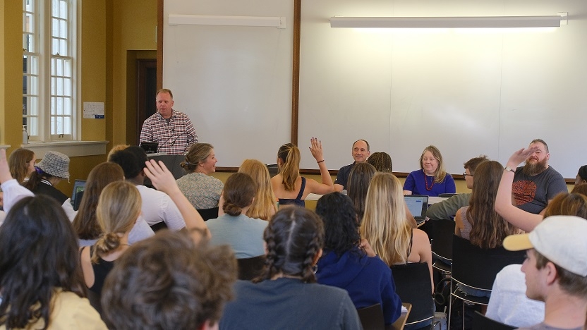 Matt Lawrence stands at the front of seated group of students. At the front of the room, three people sit facing the crowd for a panel discussion.