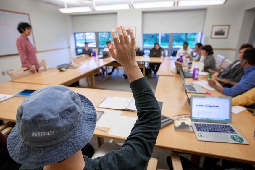 photo of middlebury class with tables set up in a square formation and a student with their back to the camera and hand raised to participate