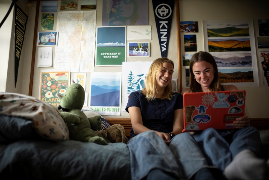 two students sit on dorm room bed with flags and posters in the background looking at a laptop