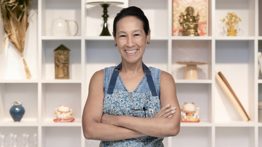 Wearing a blue shirt and apron, Robynne Maii poses for a headshot in front of white bookshelves.