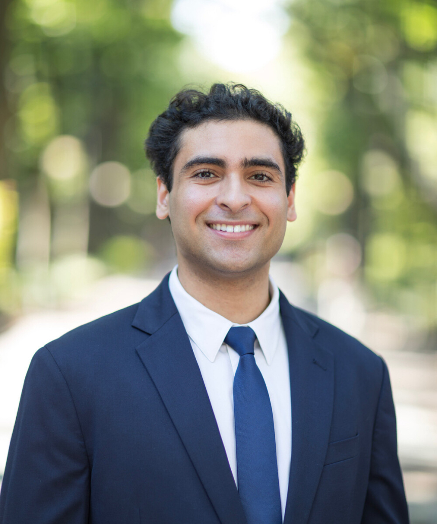 A person looks at the camera and is smiling. They are wearing a suit and a blue tie.
