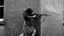 black and white photo of young woman aiming an assault rifle around the corner of a building
