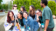 A group of 8 friends from the class of 2020 laughing while arranging themselves for a group photo. Several of them carry Middlebury tote bags featuring a cow and chapel painted by Woody Jackson.