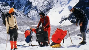 Three mountain hikers are checking their packs before they begin their climb up a large mountain in the background.